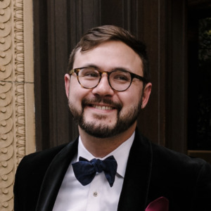 A professional portrait of Rik Jeffery. They have glasses and are wearing a tuxedo that is smiling.