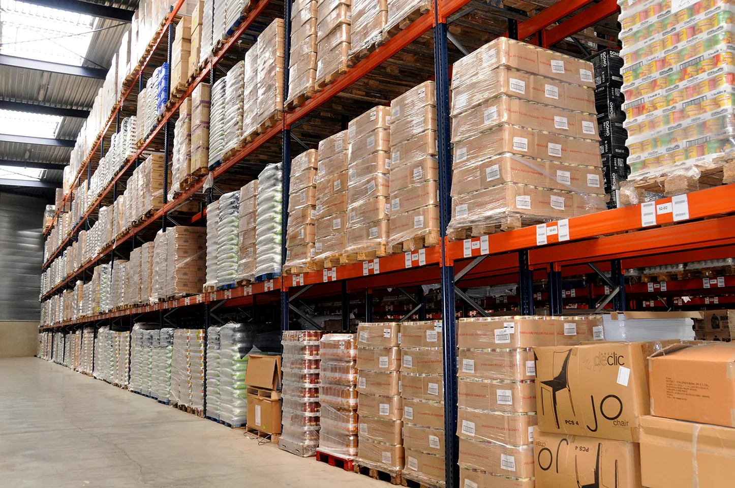 A warehouse with tall shelving units stacked with pallets of cardboard boxes and plastic-wrapped goods.