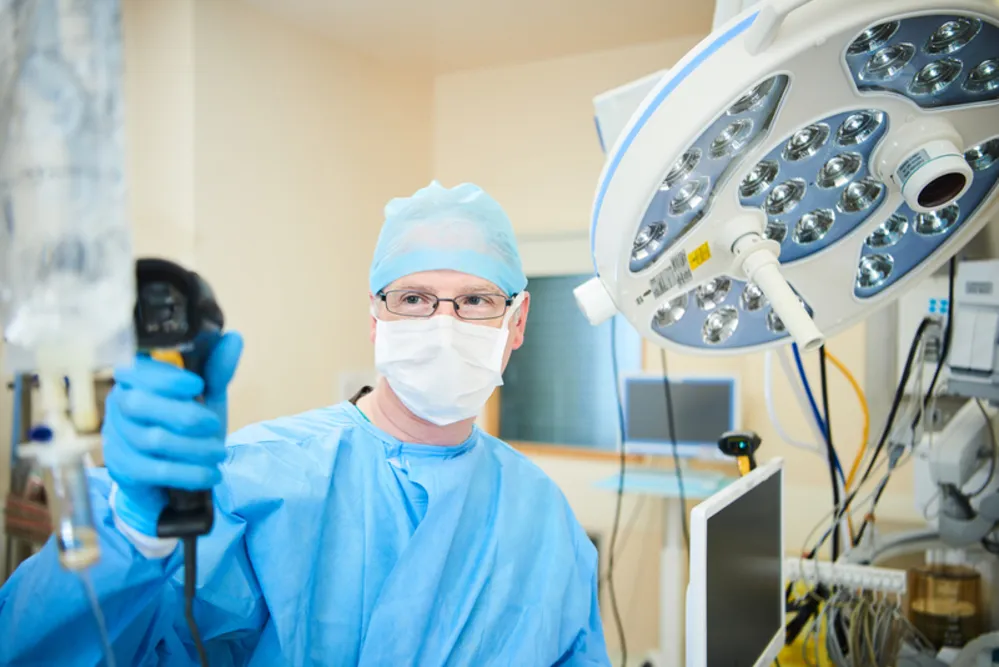 Læge iført kirurgisk kit, ansigtsmaske og handsker i et operationsrum med medicinsk udstyr.