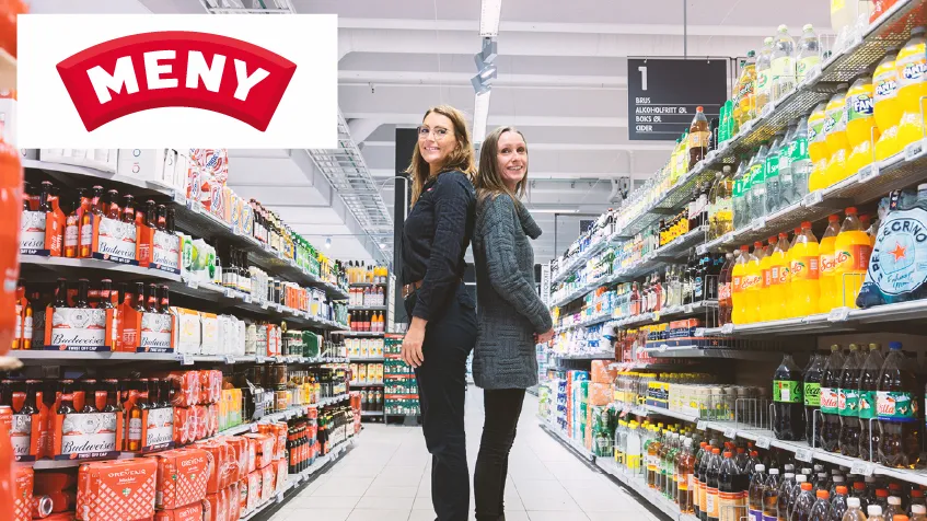 Two women are standing back-to-back in the aisle between the beverage shelves at a MENY supermarket.