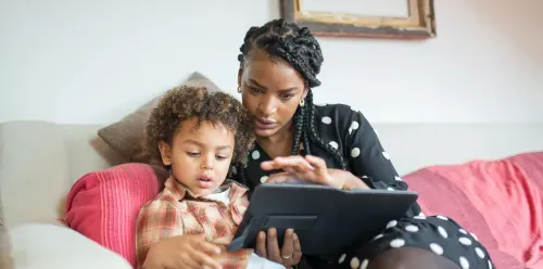 A woman and a child sitting on a couch, both focused on a tablet in their hands, sharing a moment of engagement.