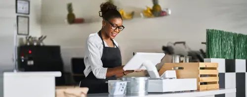 A women wearing glasses and an apron is smiling while using a touchscreen in a modern cafe setting