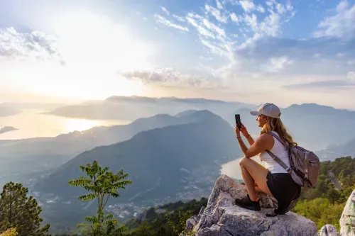 A woman with a backpack sits on a rock, focused on her phone, surrounded by nature.