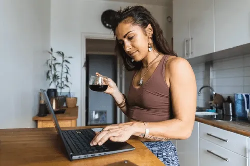 Woman standing at kitchen counter typing on a laptop while holding a glass cup of coffee.