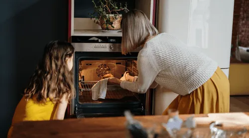 Woman using oven mitts to take a tray of baked food out of the oven while a young girl watches.