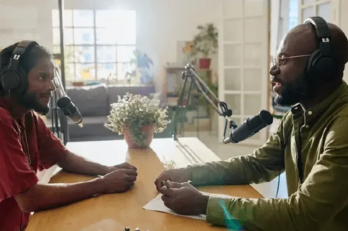 Two men wearing headphones speaking into microphones while recording a podcast in a bright room.