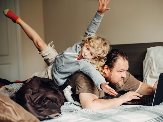 Child playfully lying on adult's back while adult uses laptop on bed.
