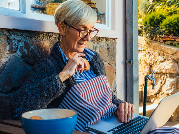Smiling older woman wearing glasses and apron working on a laptop while holding a cookie near a stone wall.