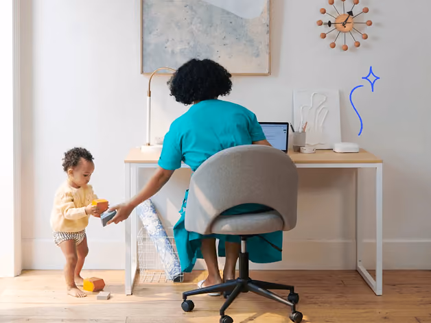 Child playing with blocks beside adult working on laptop at home desk with wall clock and artwork.