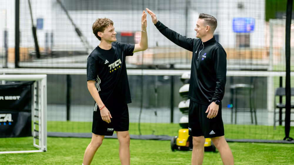 Soccer player and coach sharing a high-five during a private training session, with a TOCA technology-enabled goal and Touch Trainer system visible in the background on an indoor soccer field.