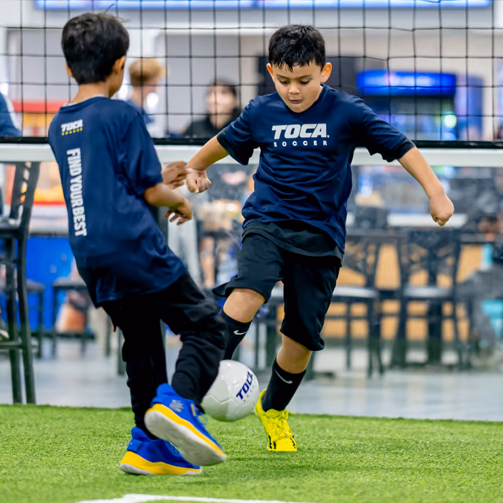 Two young players in TOCA Soccer jerseys competing for the ball on indoor turf at a TOCA Soccer facility