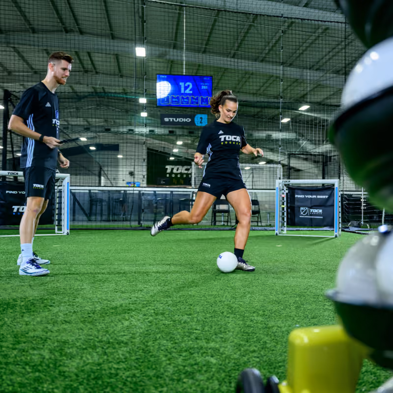 Woman kicks soccer ball on artificial turf while coach trains in an indoor training facility.