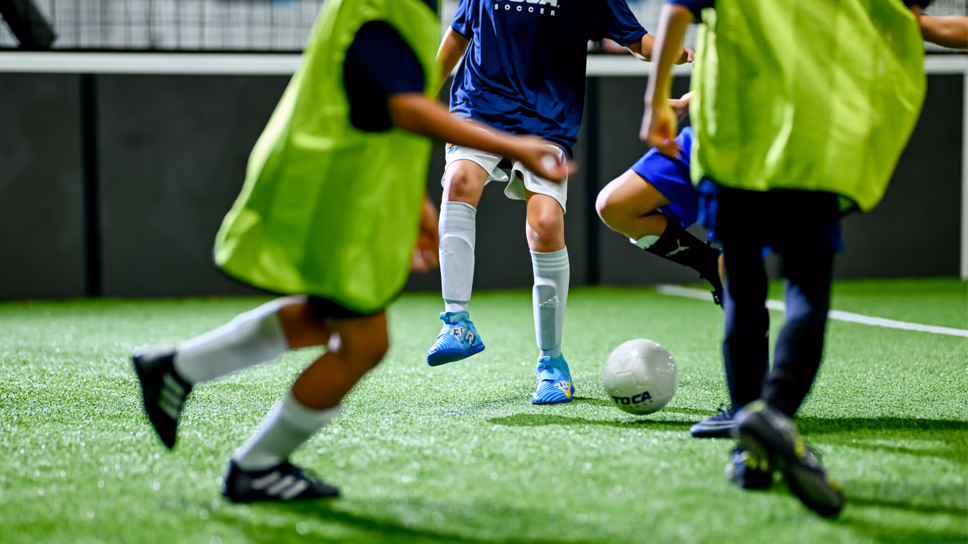 Youth soccer players in a lively pickup game at a TOCA facility, with one player about to kick the ball away from an opponent while teammates wearing pinnies look on.