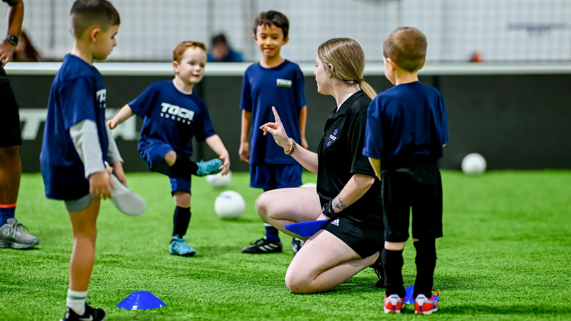 TOCA Soccer coach kneeling on indoor turf giving instructions to a group of young players during class