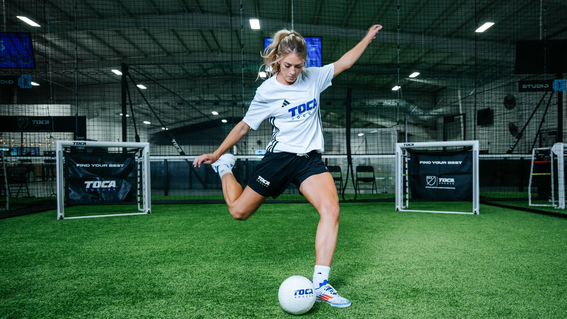 Female soccer player striking a ball on indoor turf inside a TOCA Soccer training facility