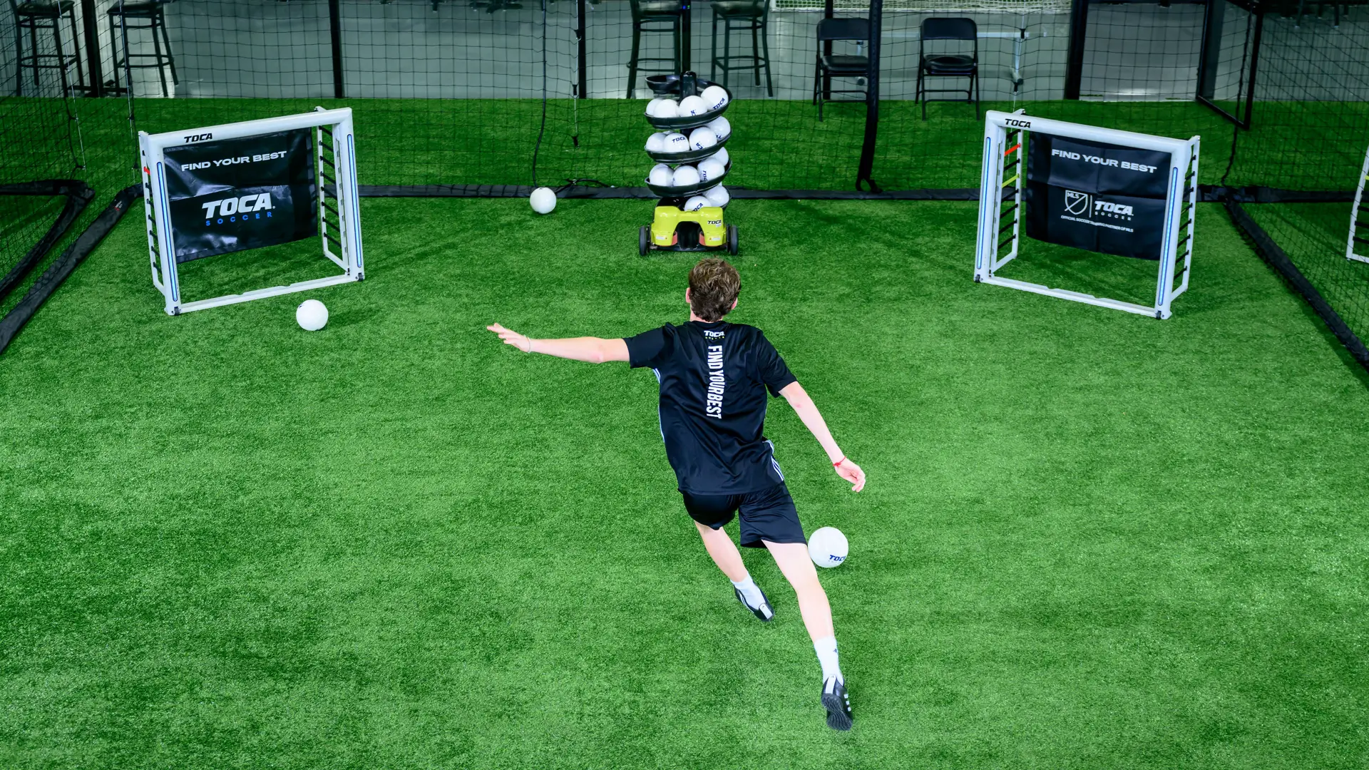 Youth soccer player shooting toward a goal on indoor turf with TOCA Touch Trainer and goals in background