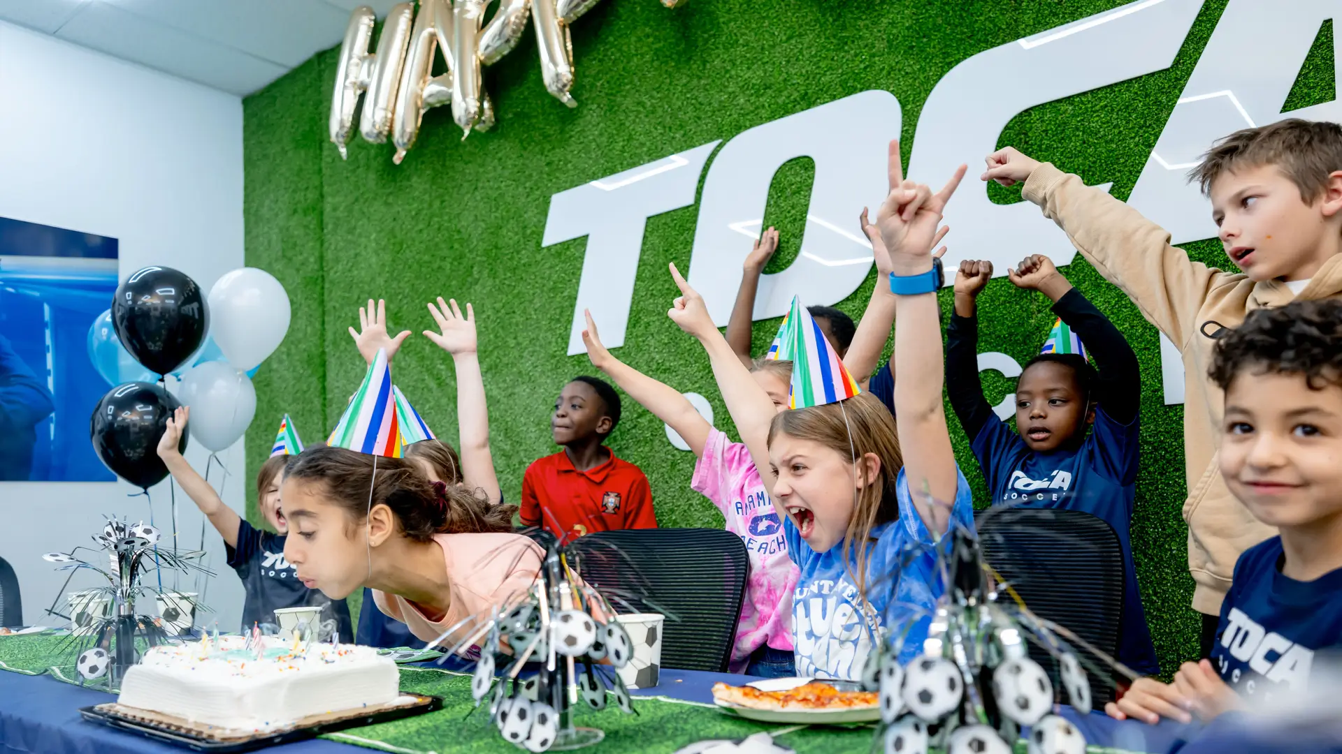 Kids celebrating a birthday party at TOCA Soccer with cake, balloons, and party hats in the party room