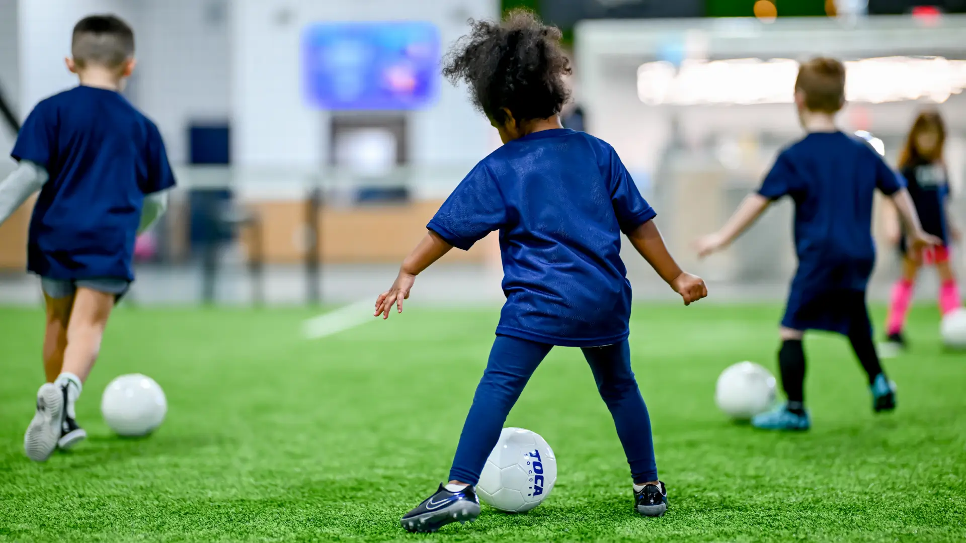 Toddlers in TOCA Soccer jerseys dribbling balls on indoor turf during a kids soccer class