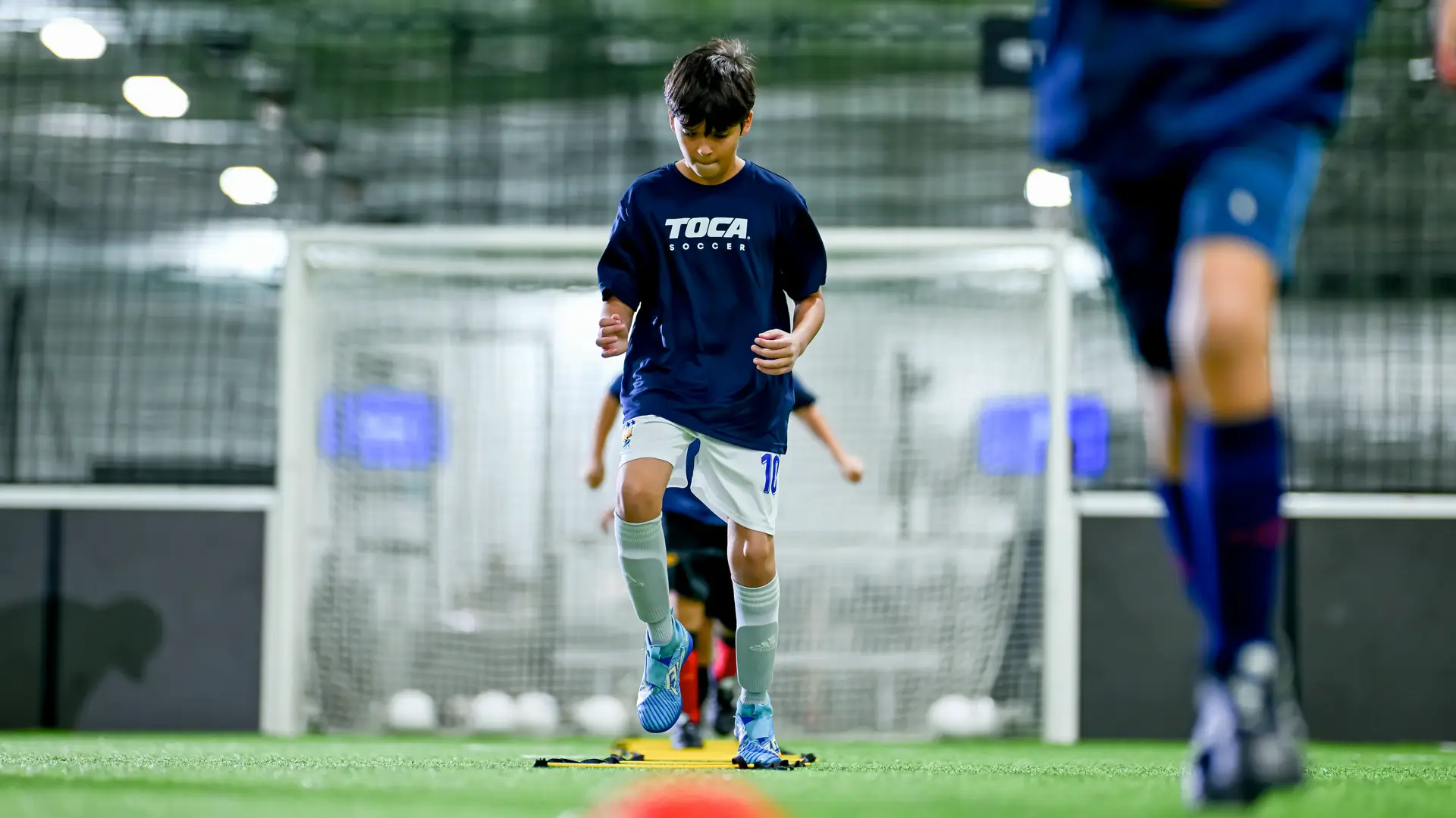 Young player in a TOCA Soccer jersey running agility ladder drills on indoor turf during training