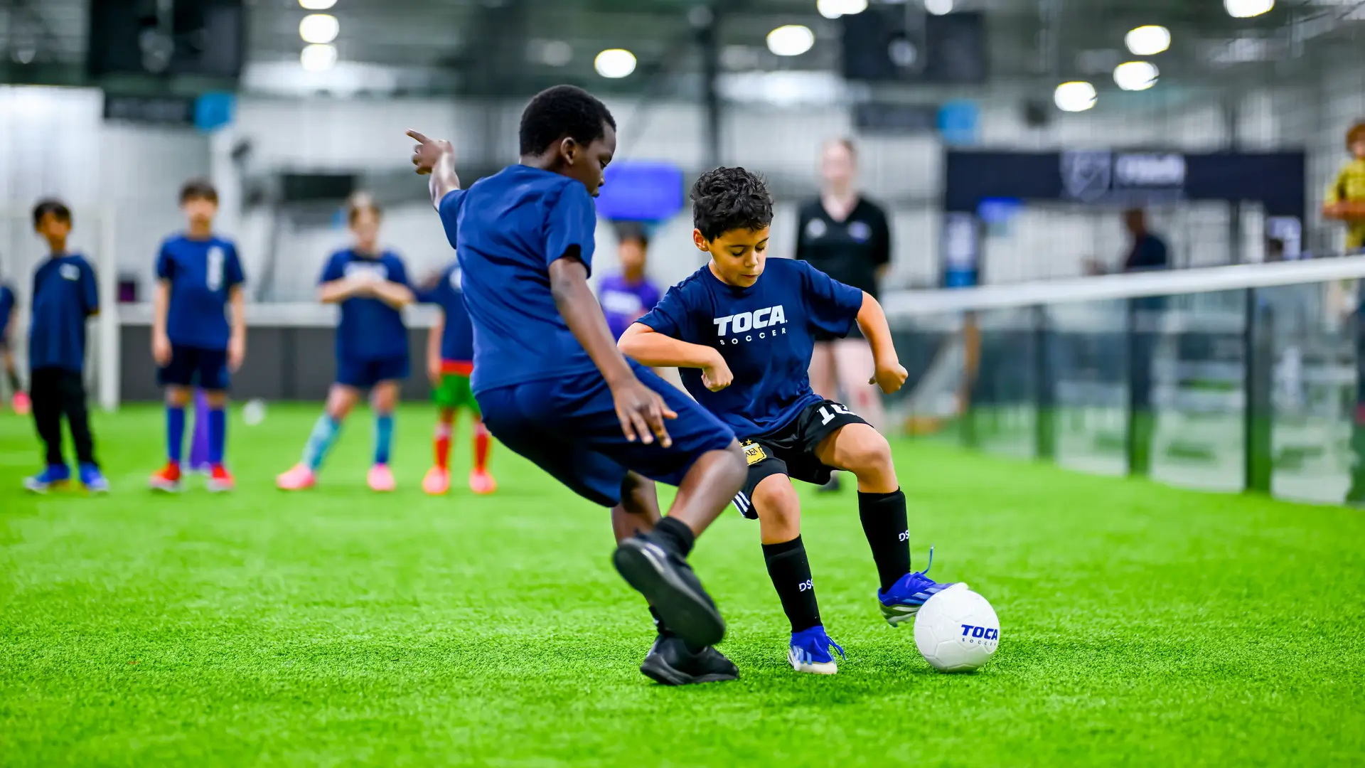 Two young players in TOCA Soccer jerseys competing for the ball during an indoor soccer drill