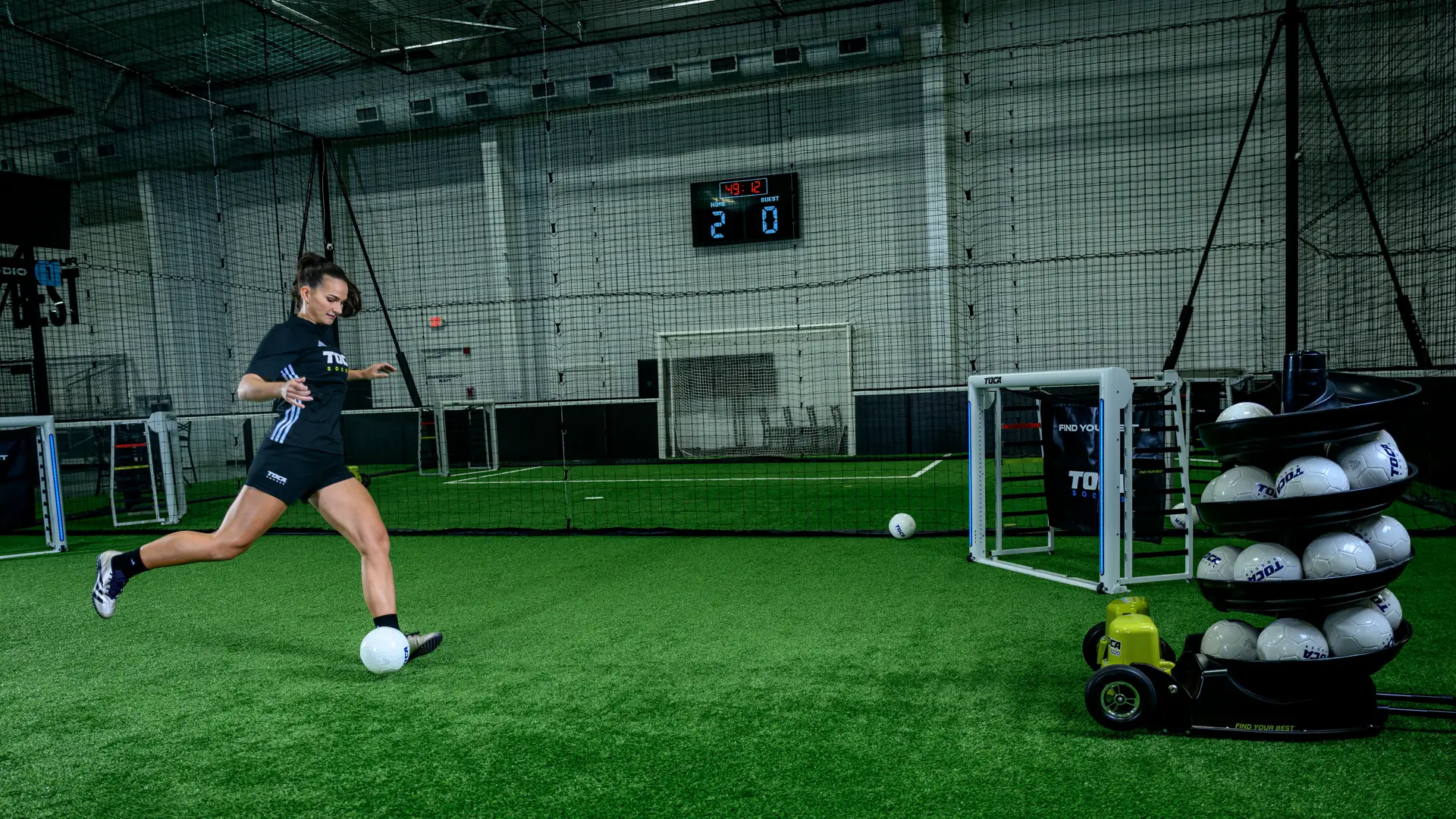 Female soccer player kicking a ball on indoor turf with TOCA Touch Trainer visible in the background