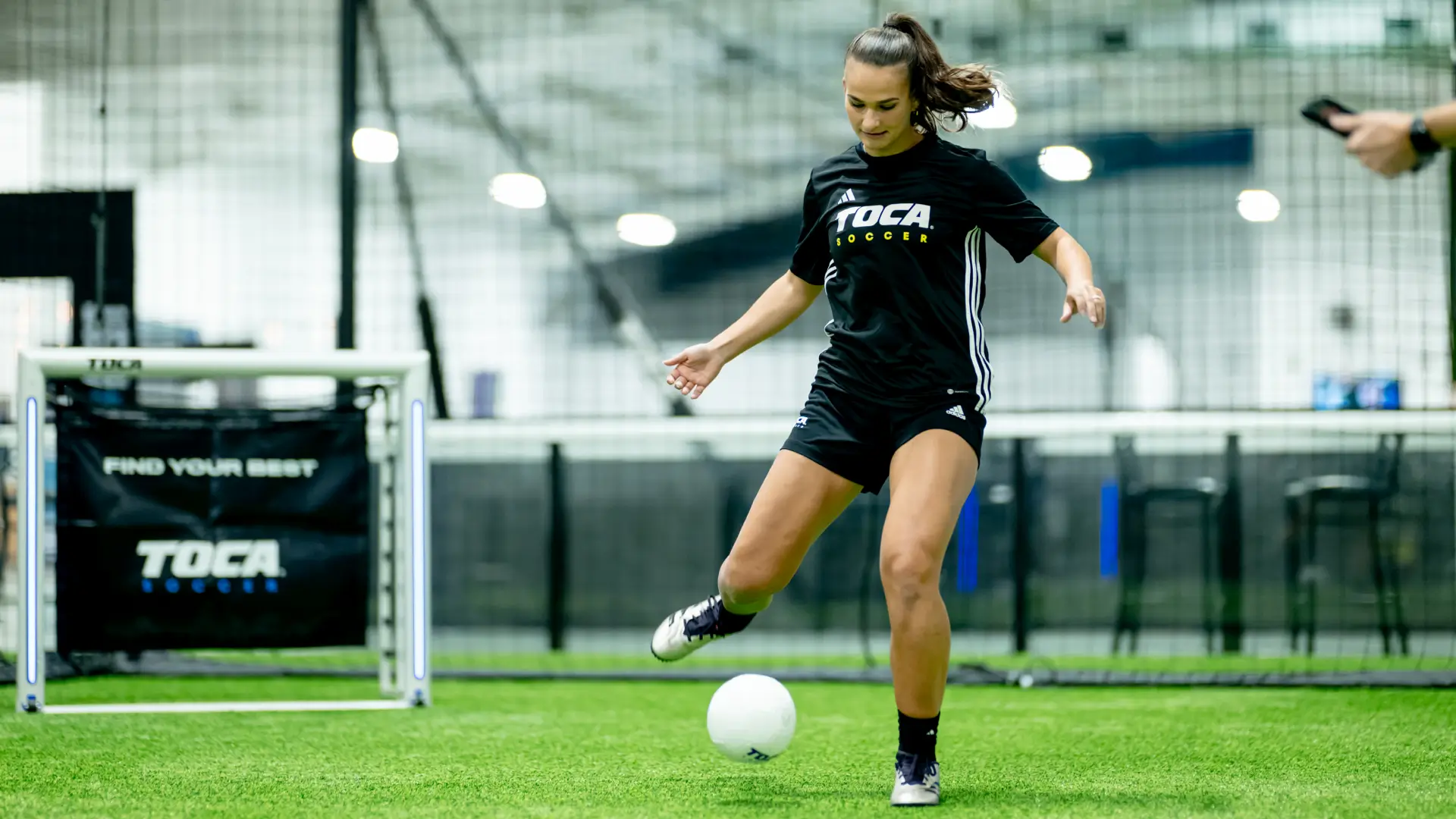 Female soccer player in a TOCA Soccer jersey about to strike a ball on indoor turf with TOCA goals and branding in the background
