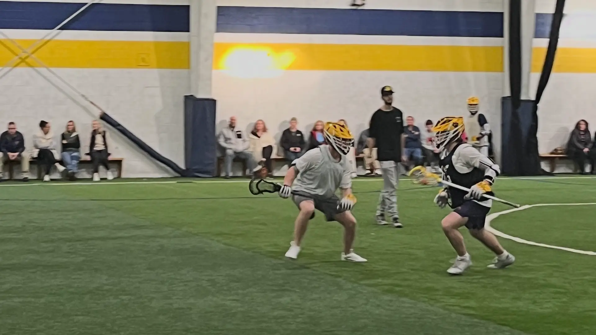 Lacrosse players competing on indoor turf with spectators watching from the sideline at a TOCA center