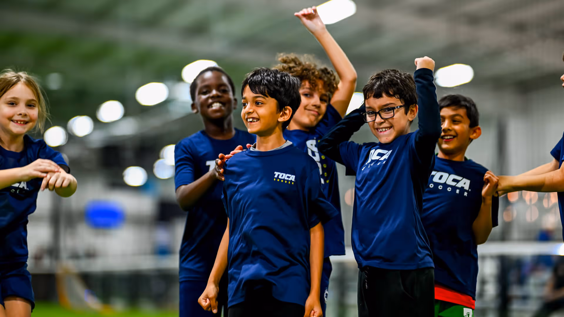 Group of young players in TOCA Soccer jerseys celebrating together with arms raised inside a TOCA Soccer facility