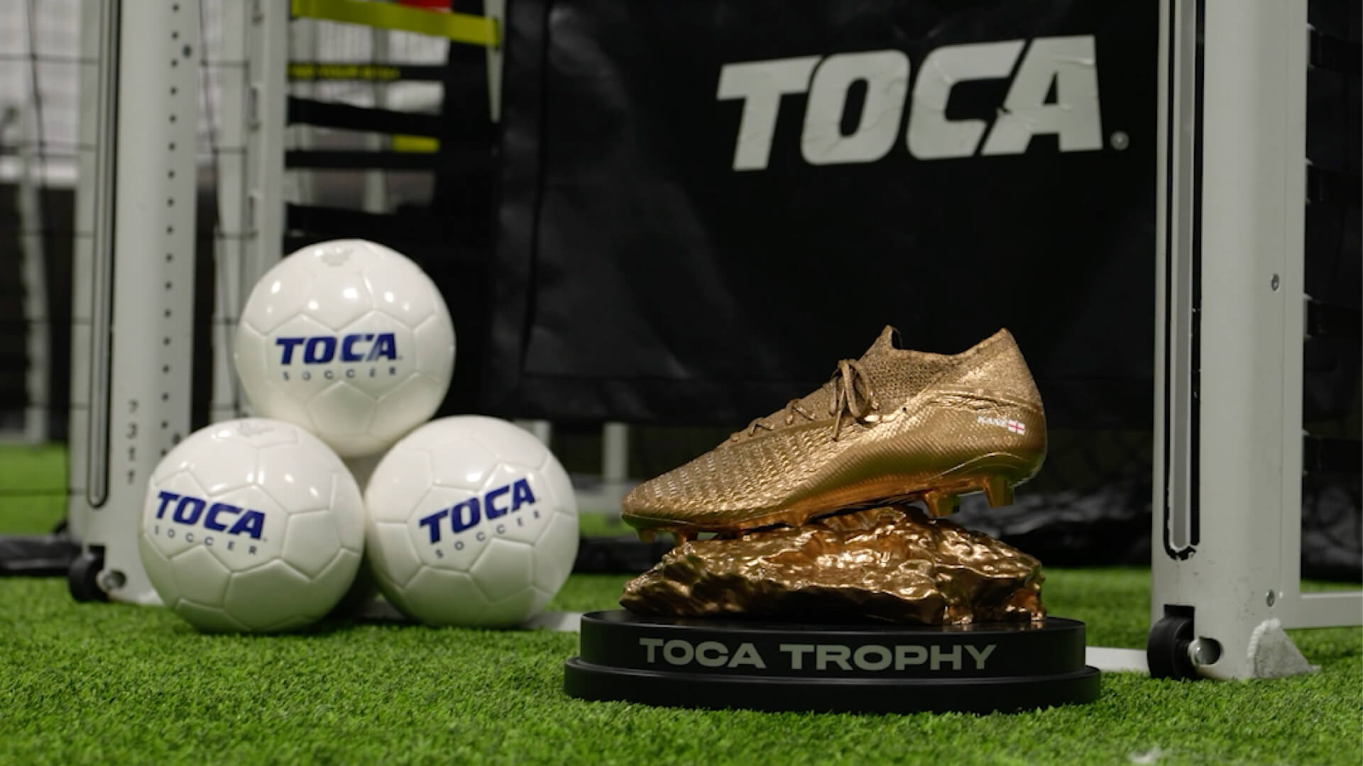 The Harry Kane Golden Boot trophy, a gold-cast soccer cleat mounted on a bronze base, displayed on indoor turf alongside TOCA Soccer branded balls at a TOCA center.
