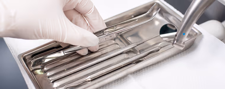 Hand in weißem Handschuh, die ein zahnärztliches Instrument aus einem Metalltablett mit verschiedenen zahnärztlichen Instrumenten aufhebt.