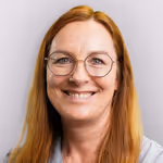 Porträt einer lächelnden Frau mit langen roten Haaren und runder Brille vor einem hellen Hintergrund.