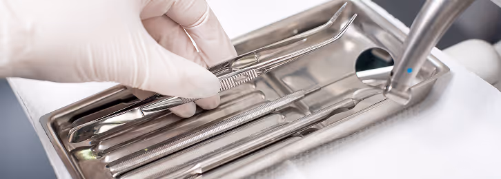 Hand mit weißem Handschuh greift nach einer zahnärztlichen Pinzette in einem steril aussehenden Metalltablett mit verschiedenen zahnärztlichen Instrumenten.