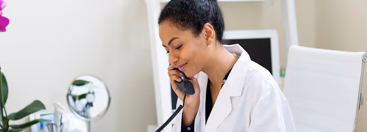 Frau in weißem Arztkittel spricht am Telefon in einem hellen Büro.