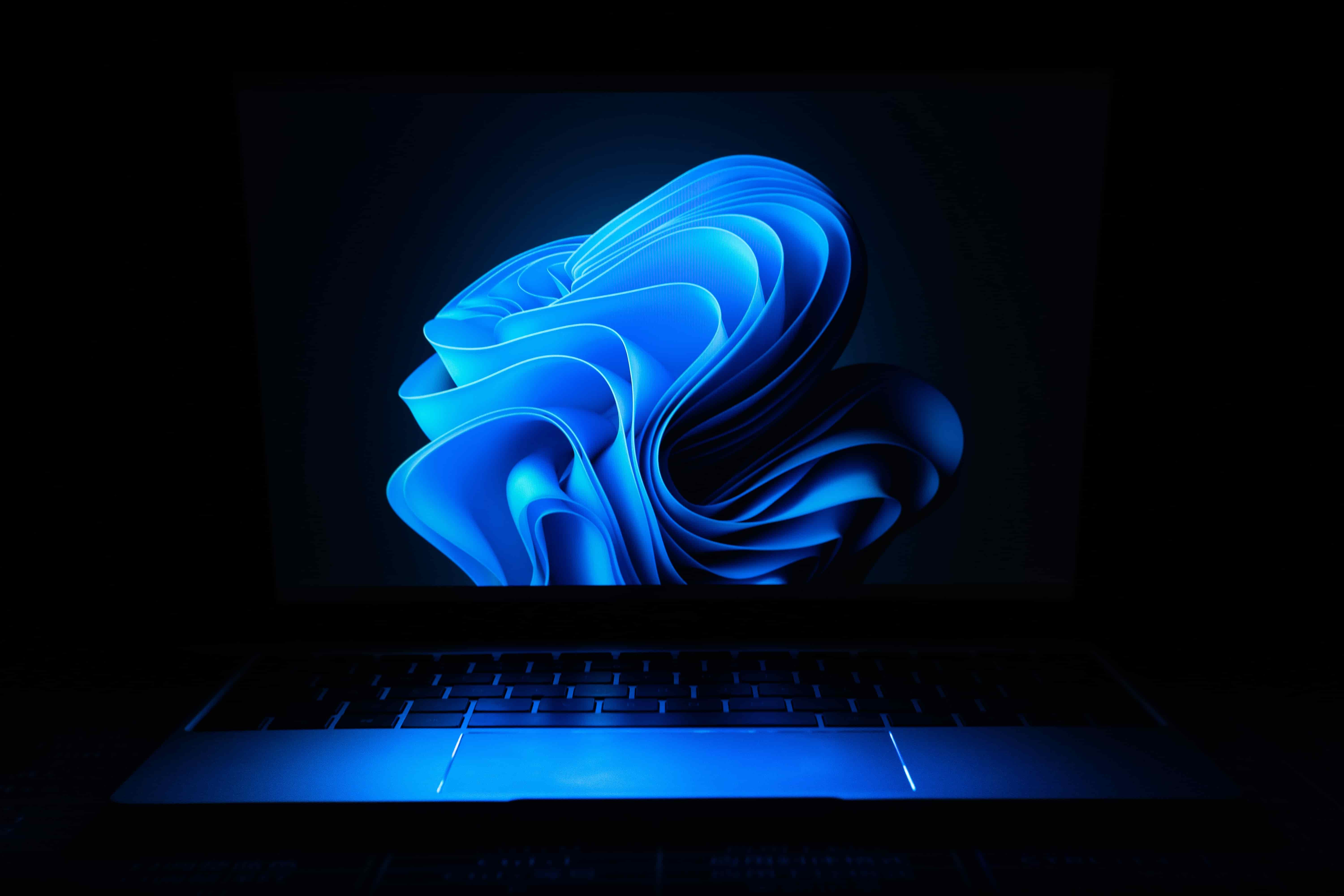 Laptop keyboard illuminated in blue light with an abstract blue swirl on the screen in a dark setting.
