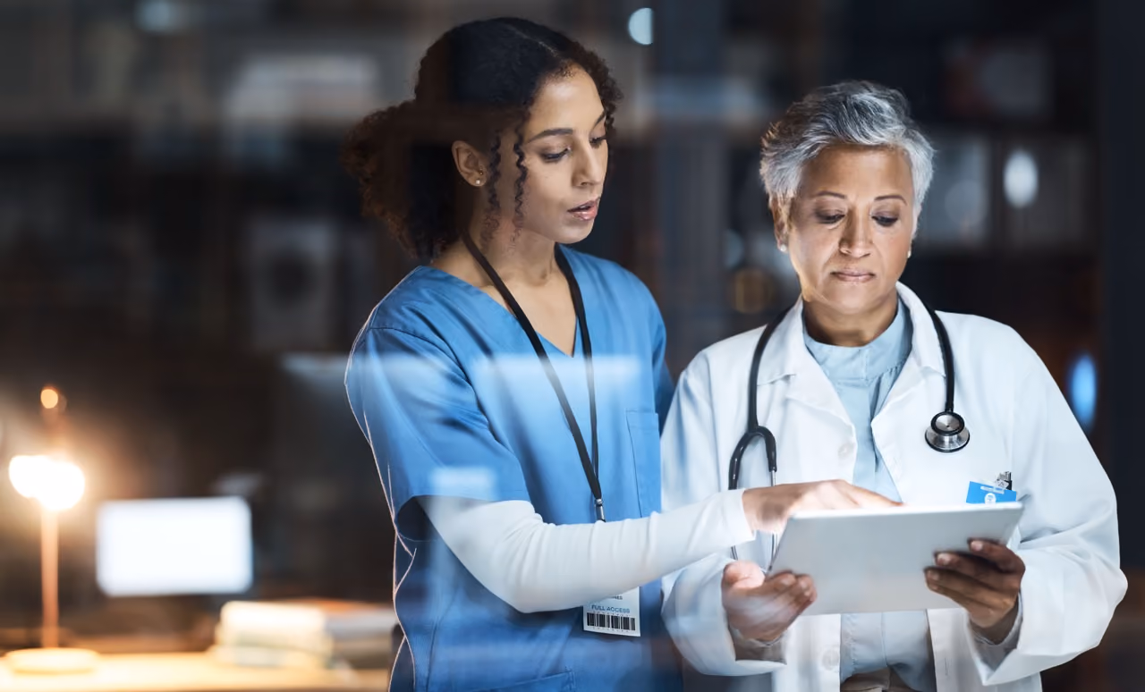 Two medical professionals reviewing information on a tablet in a dimly lit room.