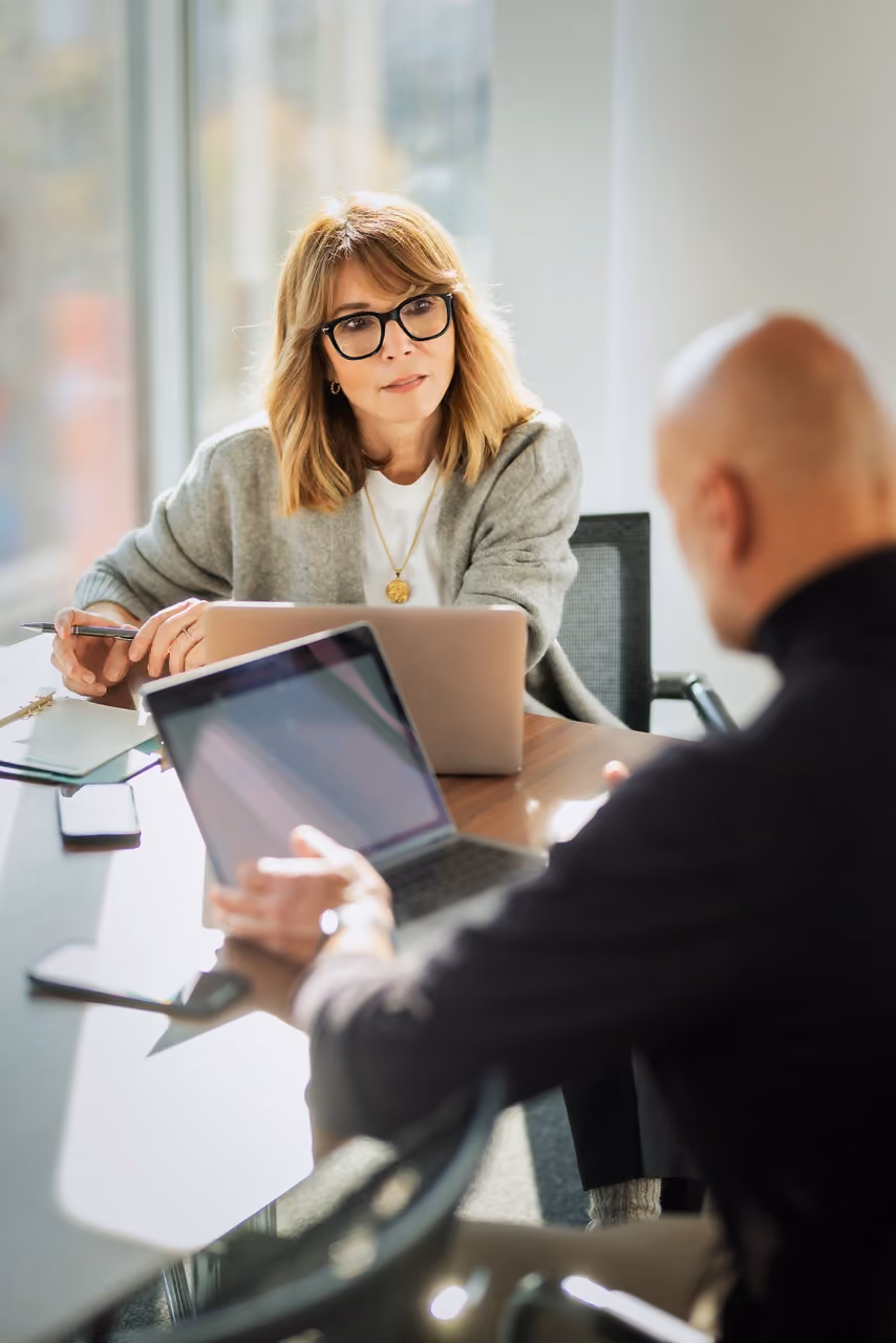 Woman with glasses and gray sweater attentively listening to man across from her in office meeting.