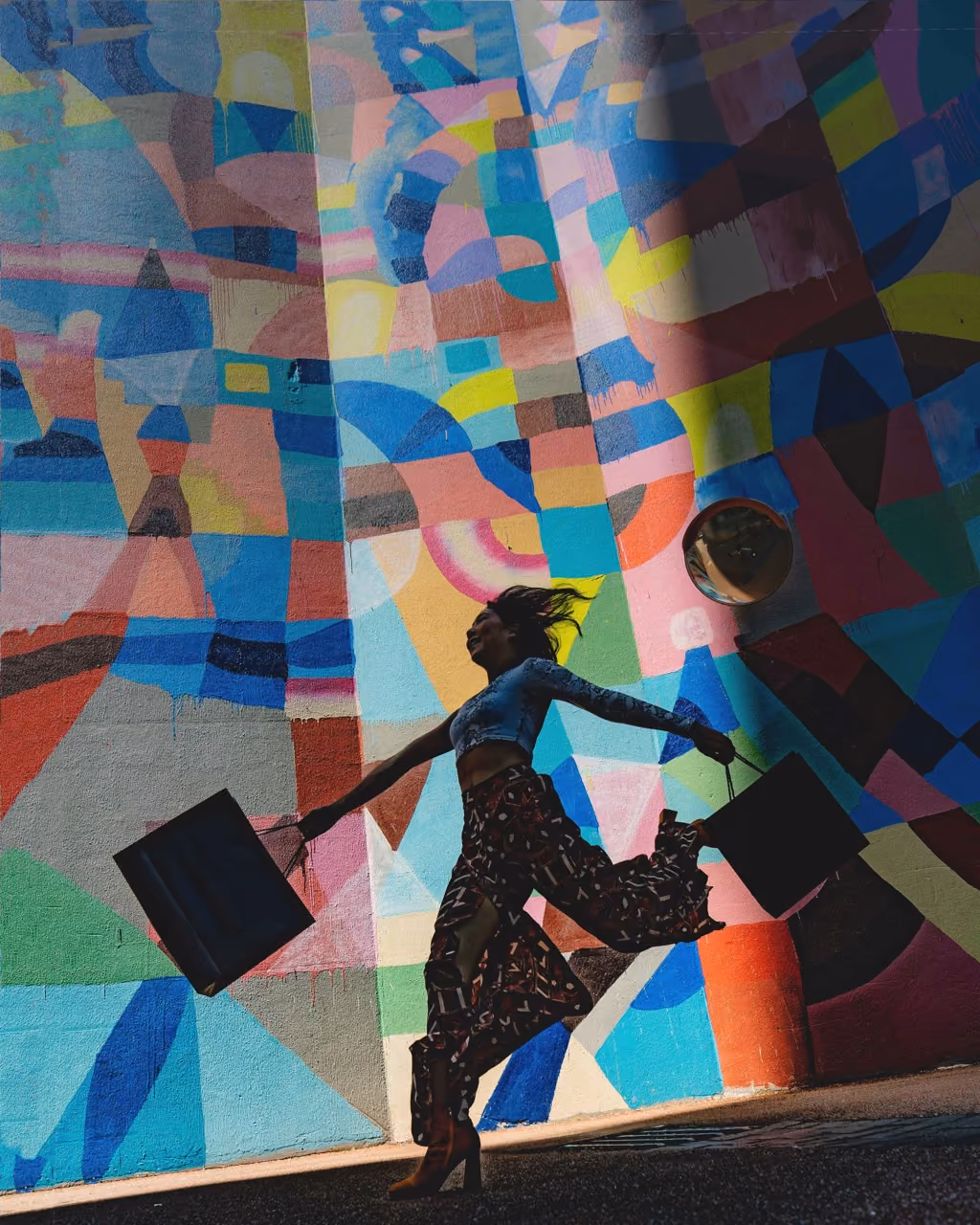 Silhouette of a woman joyfully jumping with shopping bags against colorful geometric mural.