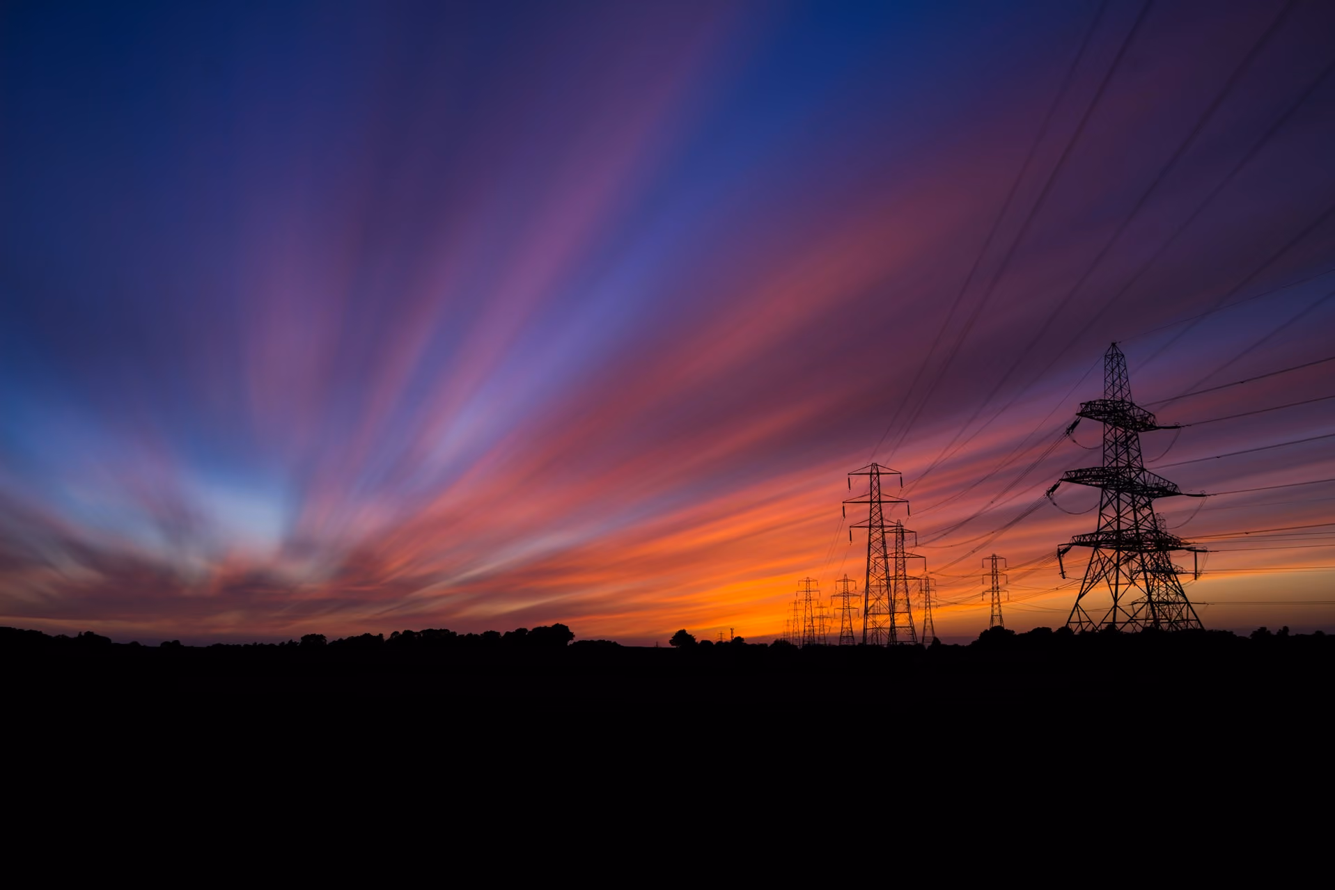 Silhouetted power lines stretch across a vibrant sunset sky with streaks of purple and orange clouds.