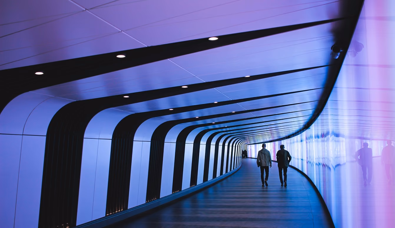 Two people walking through a futuristic blue-lit curved tunnel with black vertical supports.