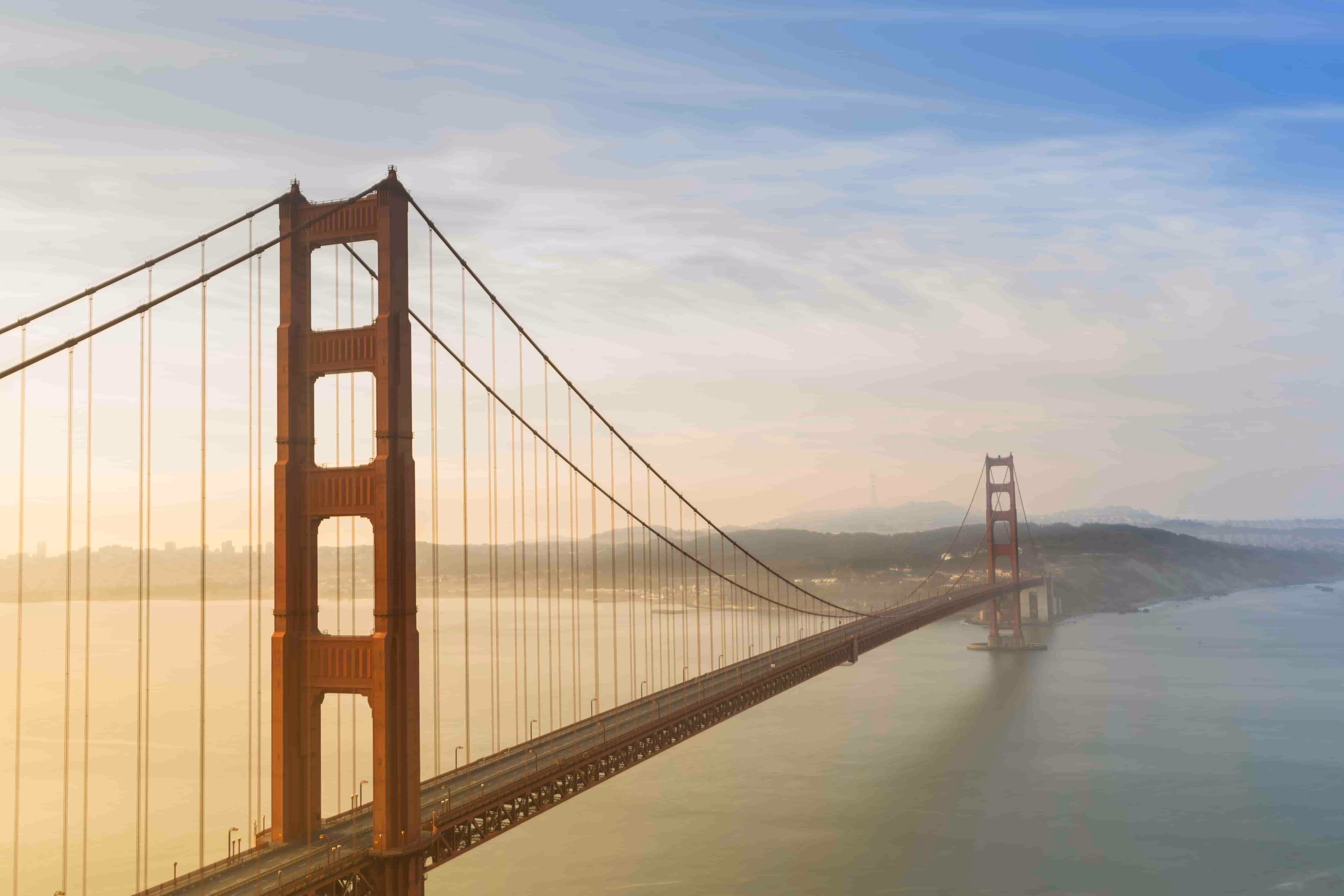 Golden Gate Bridge spanning over calm water under a partly cloudy sky at sunset or sunrise.