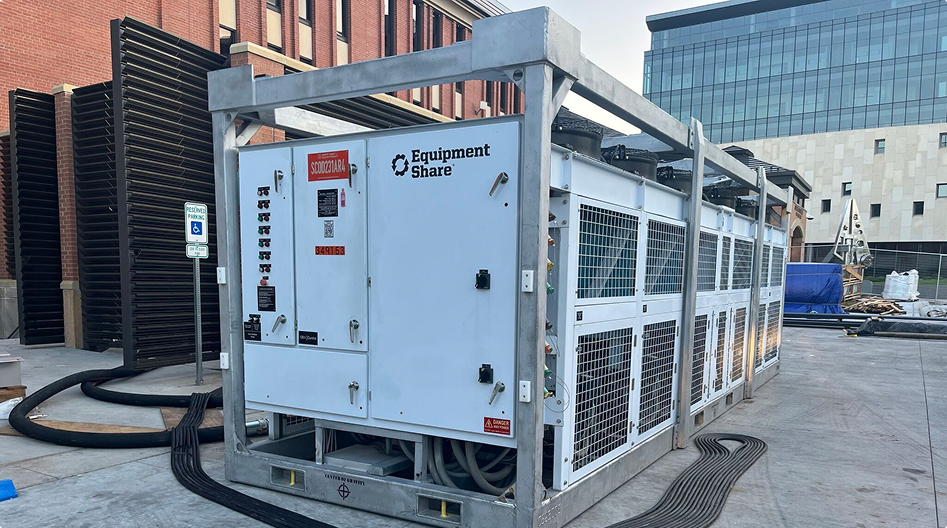 Large industrial white emergency chiller unit with Equipment Share branding, enclosed in a metal frame, sitting on a concrete ground outside buildings with hoses and cables connected to it.