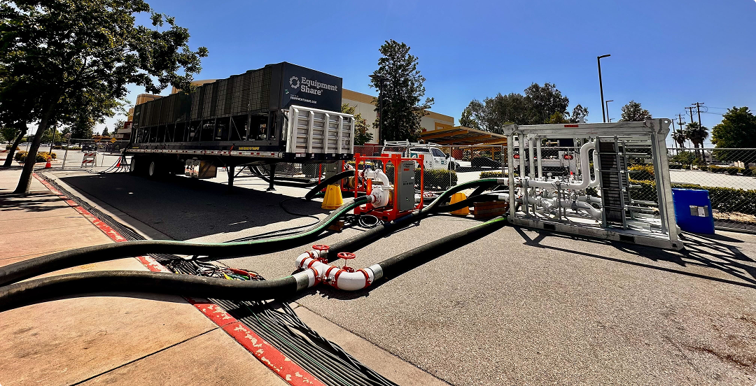 Large industrial hoses connected to a red pump system on asphalt, with a trailer labeled 'Equipment Share' and a fenced commercial area in the background under a clear blue sky.