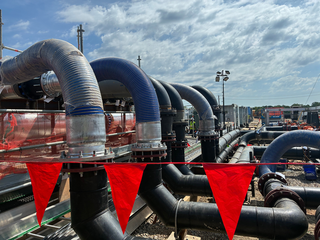 Large industrial site with multiple black and blue flexible pipes connected to metal fittings and running across a gravel ground, with red safety flags hanging in front.