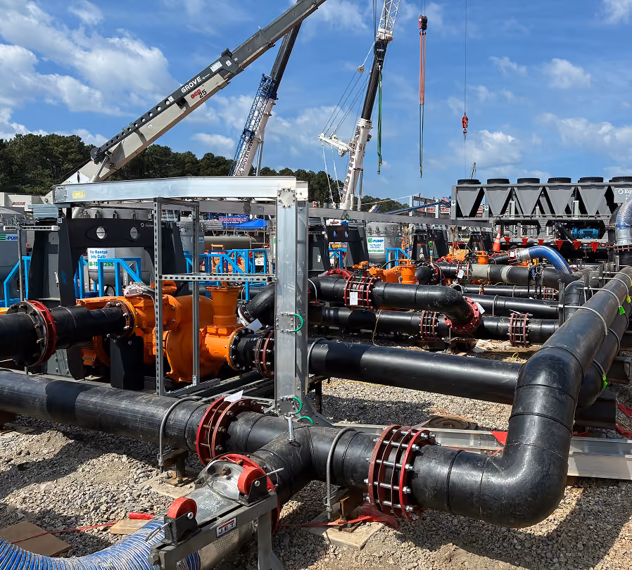 Industrial site with large black pipes connected to orange pumps and machinery, supported by metal frames on gravel ground, with cranes and blue sky in the background.