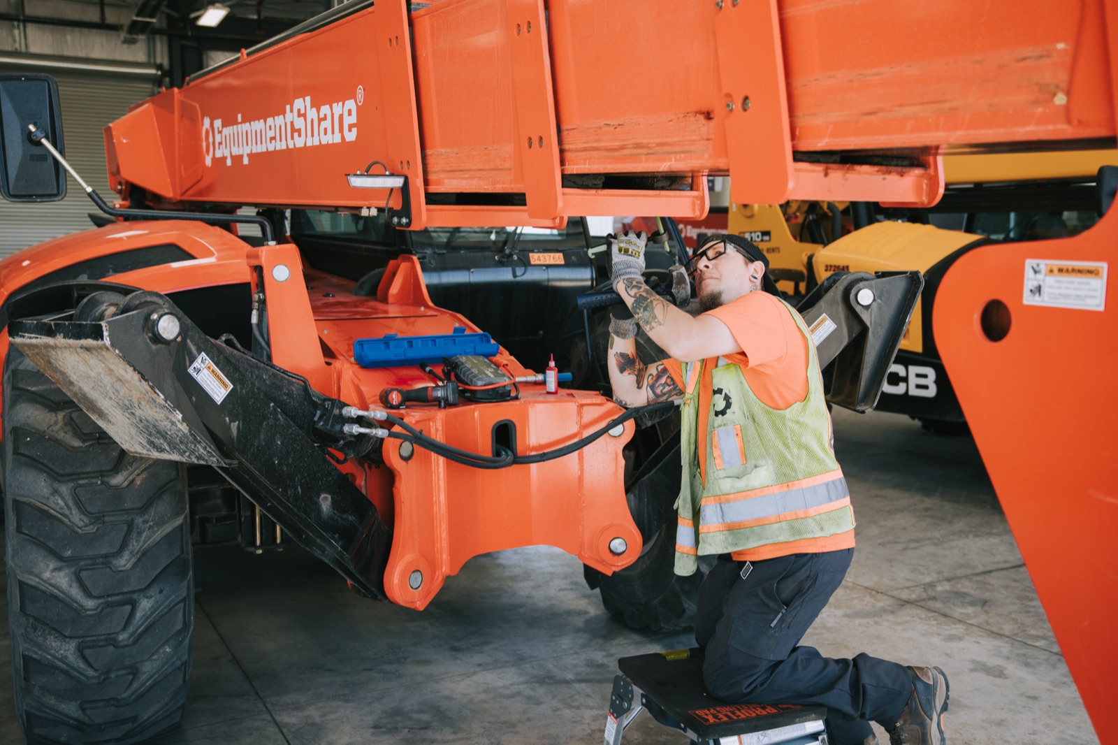 EquipmentShare technician servicing the boom of an orange telehandler in a service bay