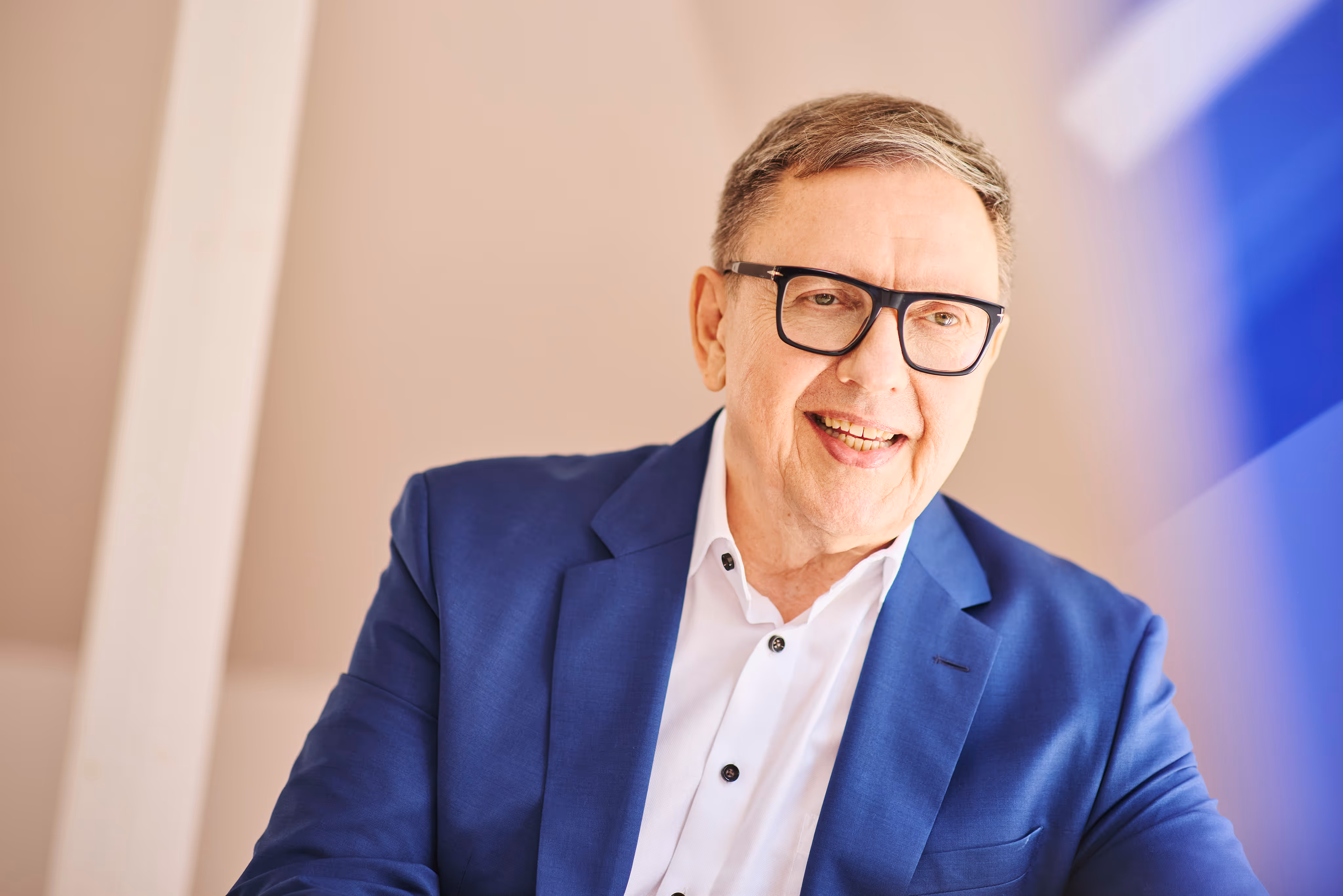 Portrait eines lächelnden Mannes mit Brille im blauen Sakko vor hellem Hintergrund