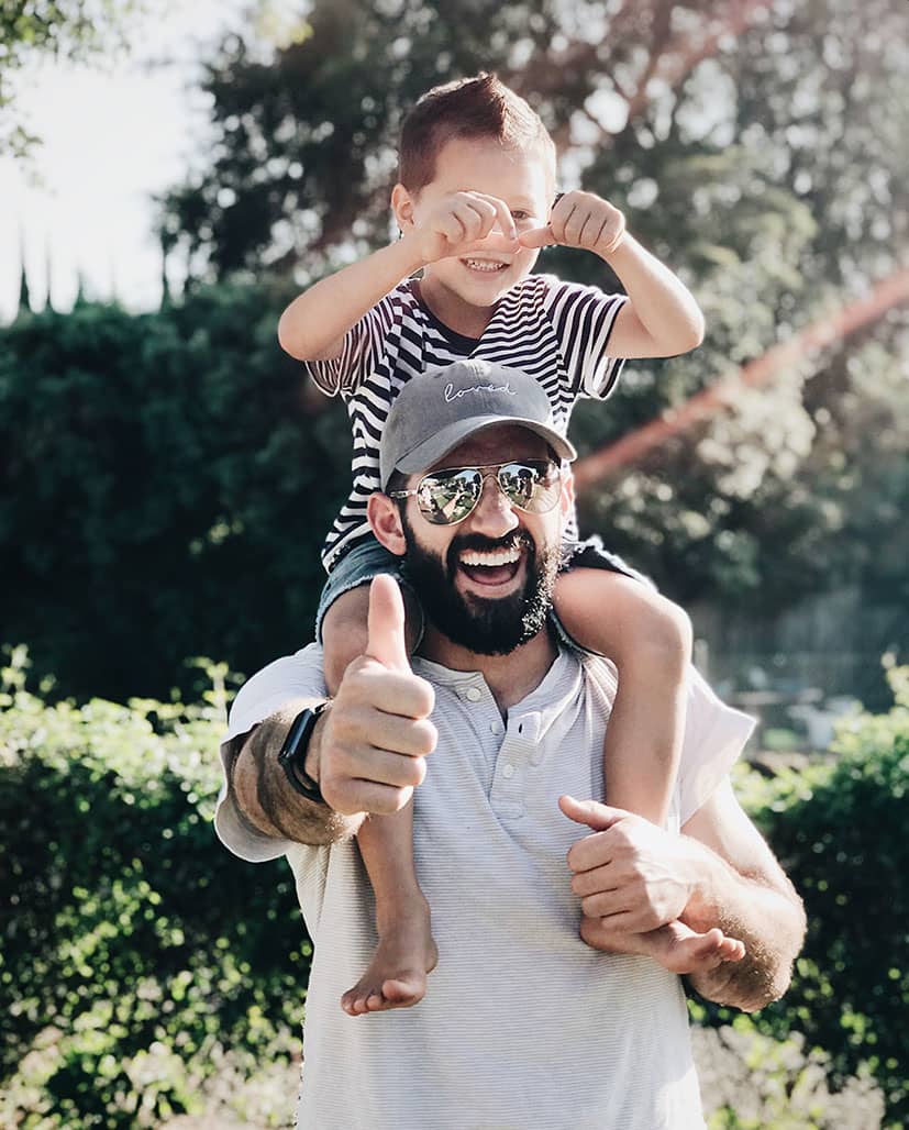 Fröhlicher Vater hält Kind auf Schultern und zeigt Daumen nach oben