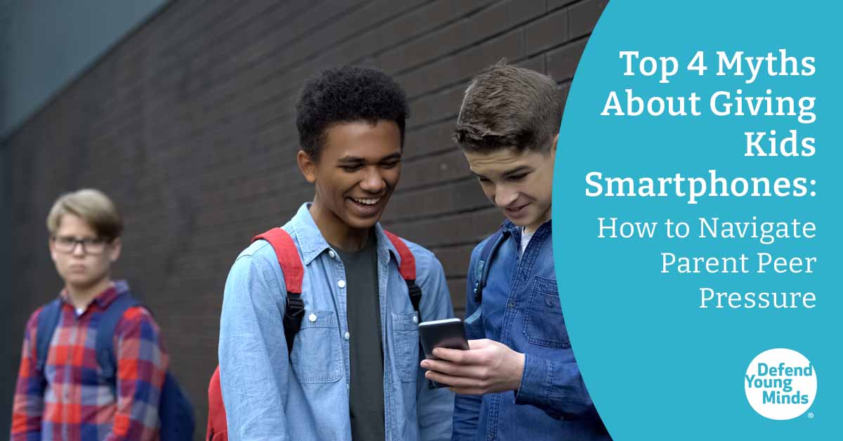Two teenagers looking at a smartphone while another teenager looks at them from a distance.