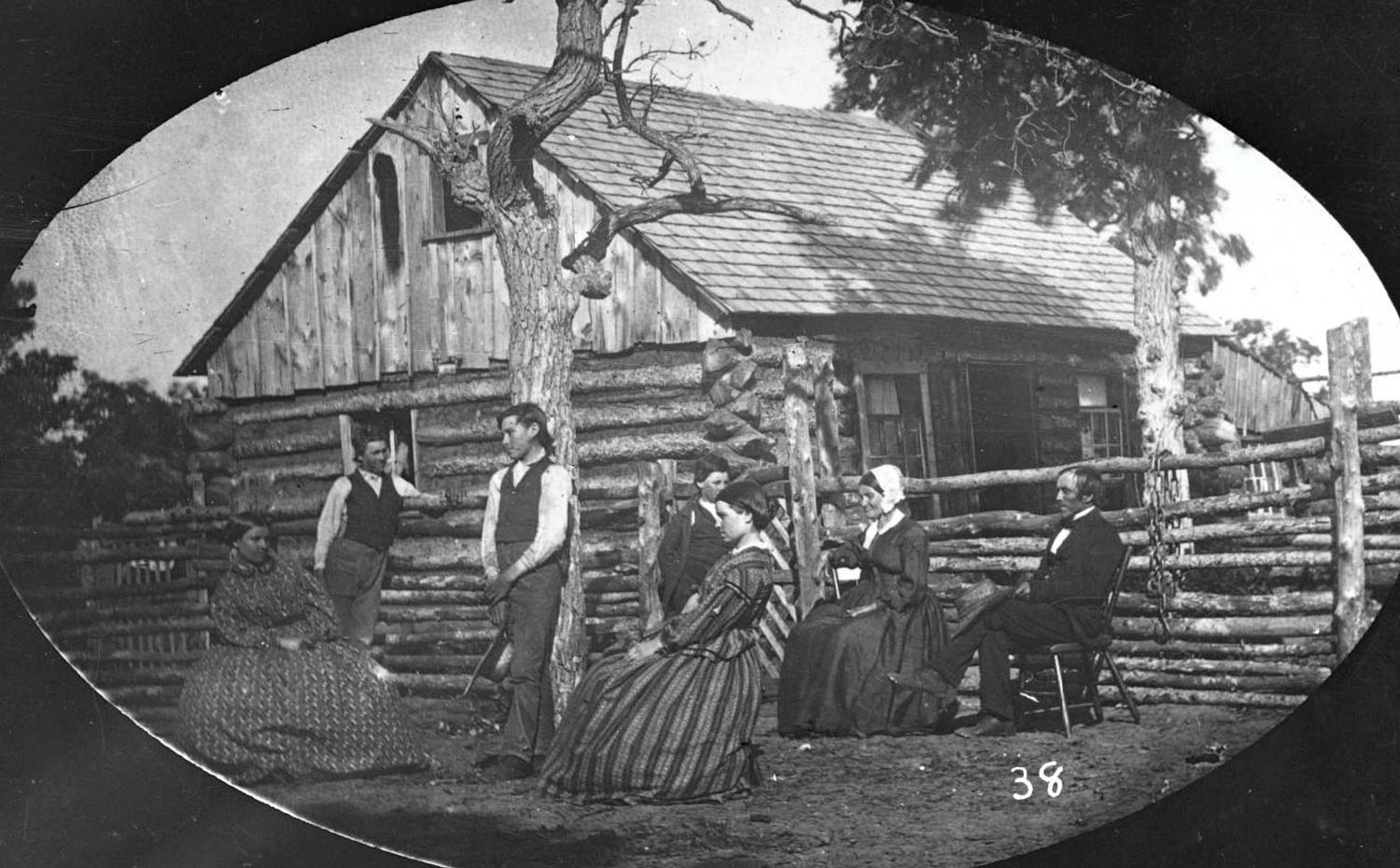 Black and white photo of a pioneer family with four women and three men seated and standing outside a log cabin with wooden fencing.