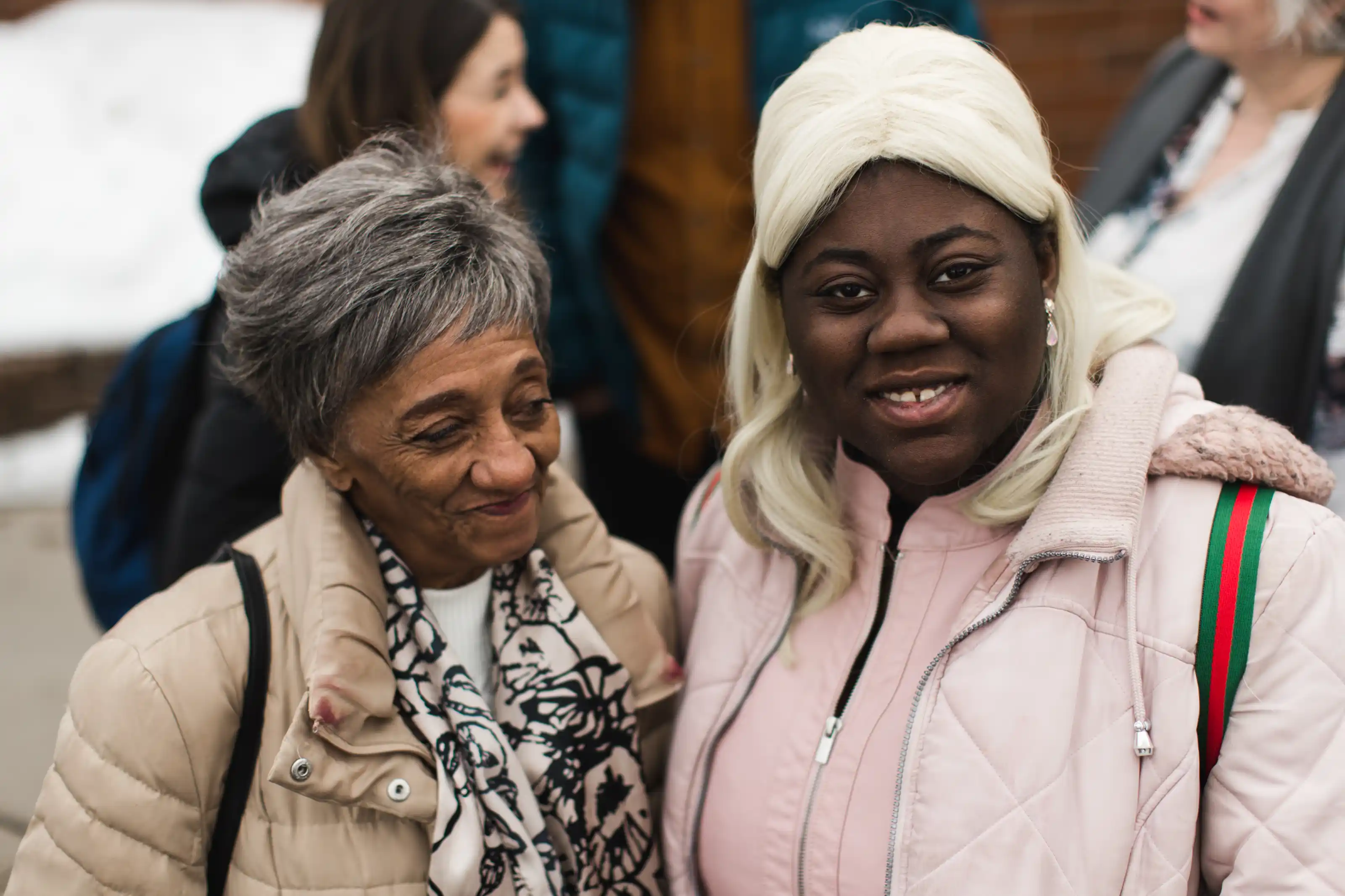 An older woman with short gray hair wearing a beige coat and patterned scarf stands next to a younger woman with long blonde hair wearing a pink jacket and a backpack.
